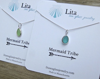 May include: Two silver necklaces featuring small, smooth, rounded sea glass pendants in light green and teal. Each necklace is displayed on a white card printed with the text Lita sea glass jewelry, Mermaid Tribe, and a black arrow graphic.