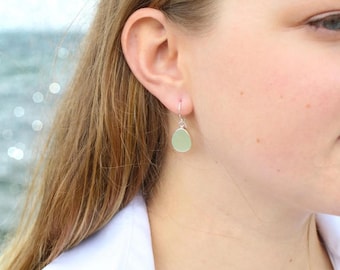 May include: A drop earring featuring a smooth, pale green piece of sea glass encased in a sterling silver setting. The earring hangs from a thin sterling silver hook. The text "Real Sea Glass . Sterling Silver ." is printed at the bottom.