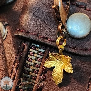 May include: A gold-colored leaf charm attached to a brown leather bag with a zipper and a metal clasp. The charm is hanging from a silver chain.