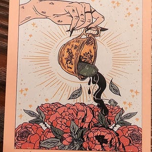 May include: A tarot card illustration featuring a hand pouring tea from a decorated cup onto a bed of red roses. The card is peach-colored with the text "THE TEA" at the bottom and "IX" at the top. The Copper Lotus Art Studio logo is in the bottom left corner.