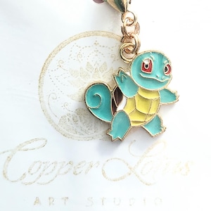 Puede incluir: Un dije dorado con un personaje de dibujos animados de Squirtle con detalles de esmalte azul, amarillo y marrón. El dije está sujeto a un cierre dorado. El fondo incluye el texto "Copper Lotus Art Studio".