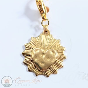 Könnte beinhalten: Goldener Anhänger mit einer Herzform, umgeben von Sonnenstrahlen. Der Anhänger hat einen Karabinerverschluss.