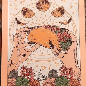 May include: A tarot card illustration featuring a taco held by hands with long nails. The card has a peach background with celestial elements, including moons and stars. The text "THE TACO" is at the bottom.