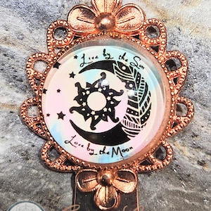 May include: Copper bookmark with a flower design. The bookmark features a round glass cabochon with a black and white illustration of a sun and moon with the text "Live by the Sun Love by the Moon".