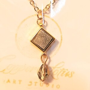 May include: A gold chain necklace with a square pendant featuring a white stone with gold trim and a dangling crystal.