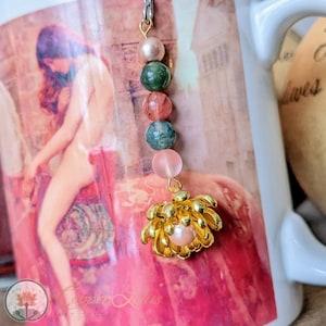 Puede incluir: Un amuleto dorado con una perla en el centro y un diseño de flor. El amuleto está unido a una cadena con cinco cuentas: una perla rosa, una piedra verde y rosa, una piedra rosa, una piedra verde y rosa y una piedra rosa.