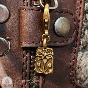 Peut inclure: Un tirette de fermeture éclair dorée avec un motif floral détaillé. La tirette est attachée à un sac en cuir brun.