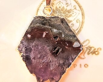 Puede incluir: Un colgante chapado en oro con un cristal de amatista en bruto de forma hexagonal. El colgante está en una cadena de plata.