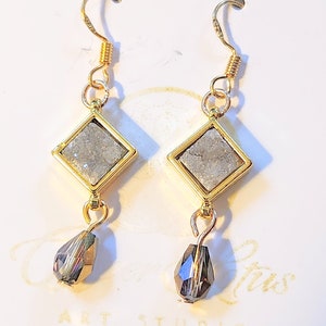 May include: Gold dangle earrings featuring a square grey stone and a teardrop-shaped black crystal. The earrings have a gold hook fastening.