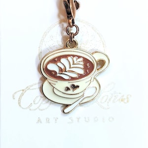 May include: A gold-tone charm featuring a white coffee cup and saucer with brown coffee and white foam art. The charm has a heart design on the saucer and a spoon. The charm is attached to a copper-tone lobster clasp.