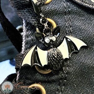 Op de afbeelding: Een zwart-witte vleermuisbedel met een goudkleurige sluiting. De vleermuis heeft een cartoonachtig ontwerp met grote ogen en een getextureerd lichaam. De vleugels zijn wit met zwarte strepen. De bedel is bevestigd aan een donkergrijze stof.