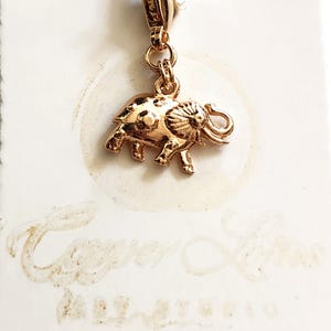 May include: A gold-colored elephant charm with a detailed design, attached to a gold-colored clasp. The charm is suspended against a white background with faint text and a circular design.