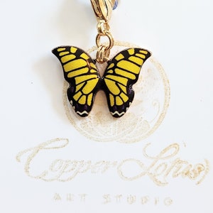 Puede incluir: Un dije dorado con un diseño de mariposa amarilla y negra. La mariposa tiene alas amarillas con contornos y detalles negros, y acentos morados. El dije está sujeto a un cierre dorado. El texto "Copper Lotus Art Studio" es visible.