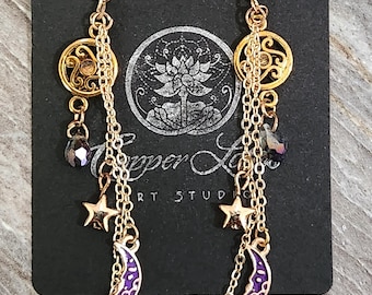 May include: A pair of gold-toned dangle earrings featuring a circular filigree charm, a small dark faceted bead, a five-pointed star charm, and a purple crescent moon charm with white filigree details, all suspended from gold-toned chains.