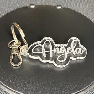 May include: A clear acrylic keychain with the name "Angela" engraved in a cursive font. The keychain has a silver metal ring and a silver metal clasp.