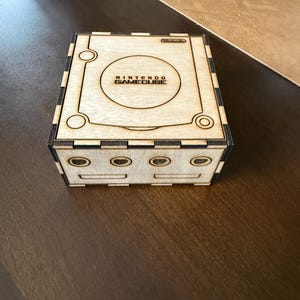 May include: A wooden box with the Nintendo GameCube logo and a cutout for the console. The box has a brown wood finish and black accents.