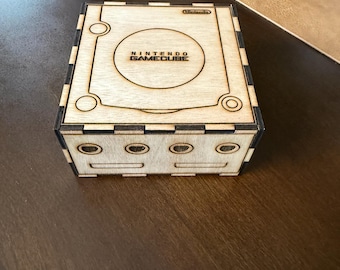 Puede incluir: Una caja de madera con el logotipo de Nintendo GameCube y un recorte para la consola. La caja tiene un acabado de madera marrón y detalles negros.