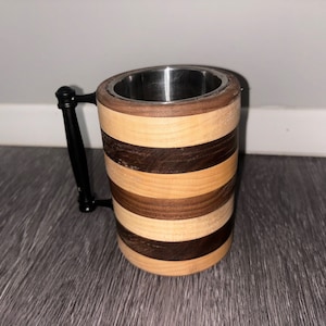 Puede incluir: Una taza de madera con un revestimiento de acero inoxidable. La taza tiene un asa negra y está hecha de madera clara y oscura alternadas.
