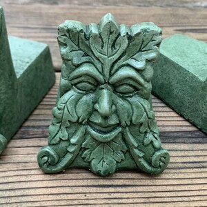 May include: A green, leafy, sculpted face with a whimsical expression. The face is made of concrete and has a textured surface.