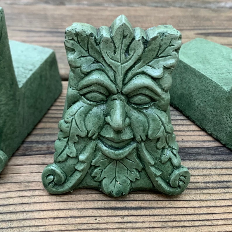 May include: A green, leafy, sculpted face with a whimsical expression. The face is made of concrete and has a textured surface.