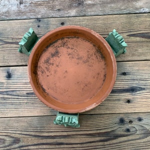 May include: A terracotta saucer with three green ceramic feet. The saucer is slightly dirty and has a rough texture.