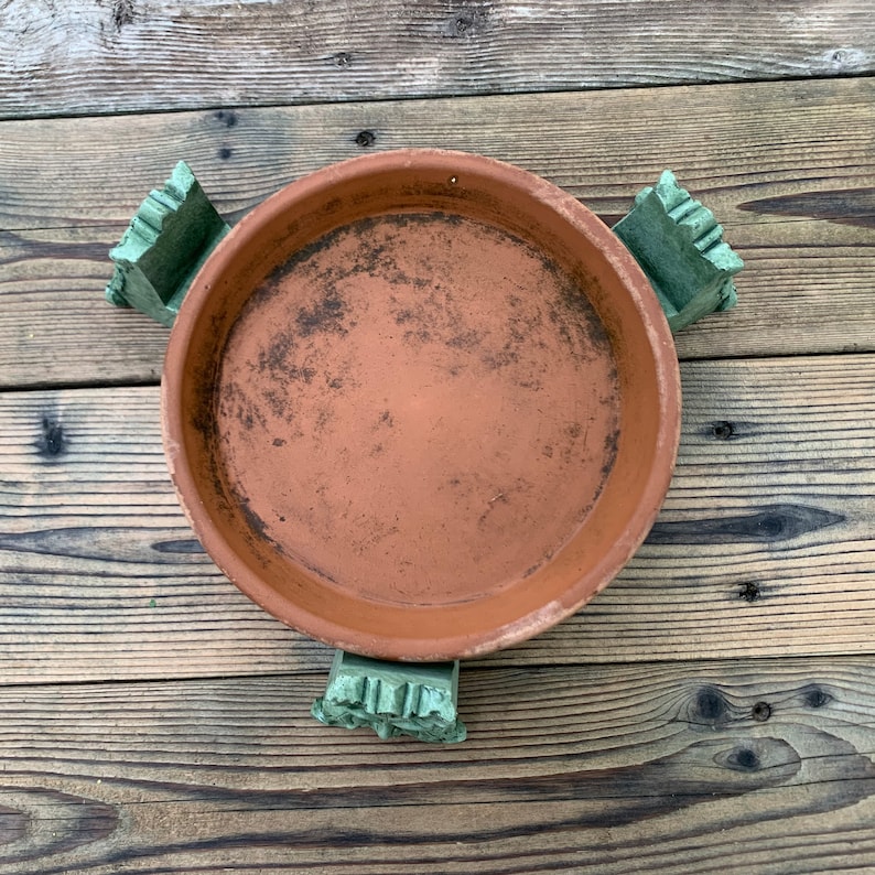 May include: A terracotta saucer with three green ceramic feet. The saucer is slightly dirty and has a rough texture.
