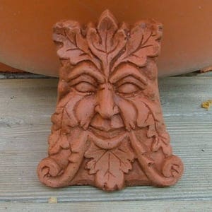 Op de afbeelding: Een terracotta groene man sculptuur met een glimlachend gezicht en bladeren gebeeldhouwd rond het gezicht en de baard.