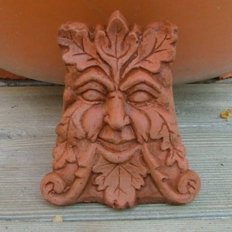 Op de afbeelding: Een terracotta groene man sculptuur met een glimlachend gezicht en bladeren gebeeldhouwd rond het gezicht en de baard.