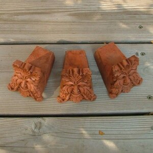 Op de afbeelding: Drie terracotta groene man sculpturen met gedetailleerde gravures van gezichten en bladeren. De sculpturen zijn in een rustieke stijl en hebben een roodbruine kleur.