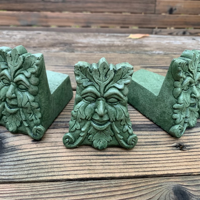 May include: Three green concrete garden ornaments shaped like a face with leaves. The face is smiling and has a whimsical expression.