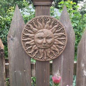 May include: A brown, wooden, sun and moon wall sculpture with a detailed face and a textured surface. The sculpture is mounted on a wooden fence.