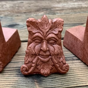 May include: A terracotta green man sculpture with a smiling face and leafy details. The sculpture is a decorative accent for a garden or home.