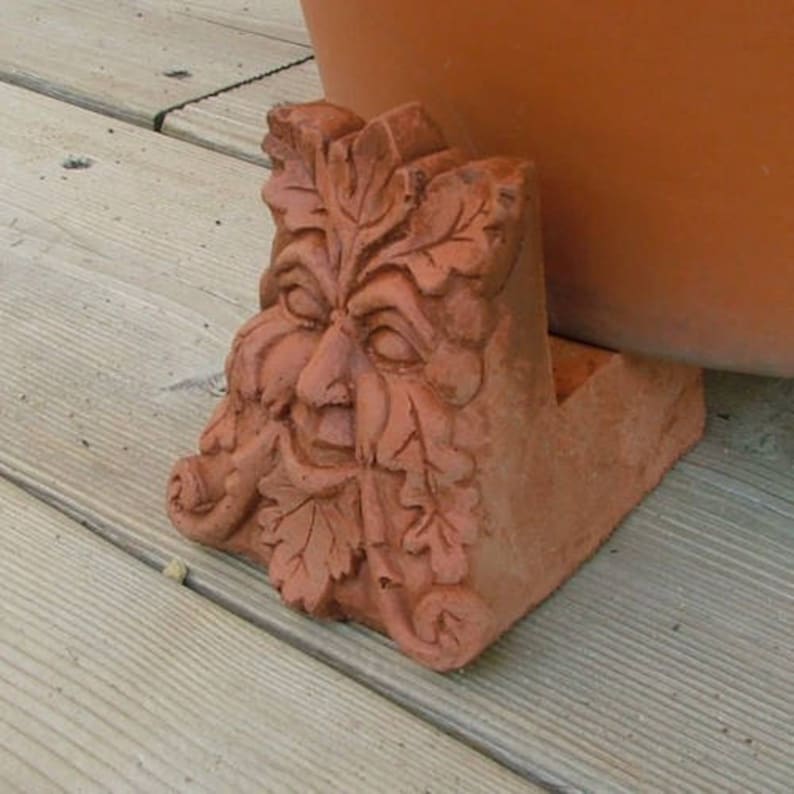 Op de afbeelding: Terracotta plantenstandaard met een groene man, een gedetailleerd gezicht en bladeren. De standaard is ontworpen om een pot te verhogen.