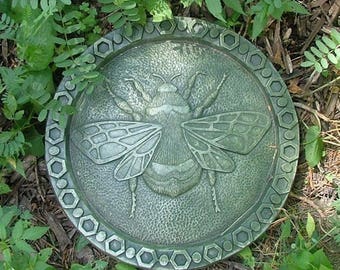 Puede incluir: Un paso de jardín de concreto verde con un diseño en relieve de una abeja. La piedra tiene un borde hexagonal.