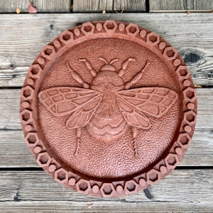 May include: A round, terracotta-colored wall plaque with a raised, detailed image of a bumblebee. The plaque has a decorative border of hexagonal shapes.