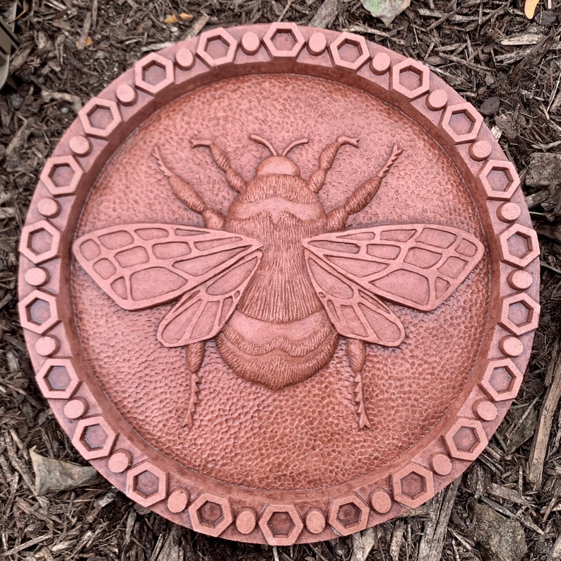 May include: A round, copper-colored wall plaque with a raised, detailed image of a bumblebee. The plaque has a decorative border of hexagons.