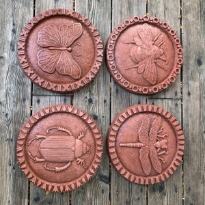May include: Four terracotta-colored round stepping stones with raised designs of a butterfly, a bee, a beetle, and a dragonfly. Each stone has a decorative border.