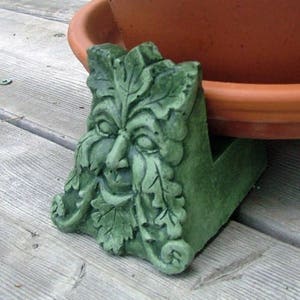 May include: Green Man planter stand made of concrete. The stand features a detailed sculpture of a face with leaves and vines. The stand is designed to elevate a planter pot.
