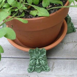May include: A terracotta flower pot with green plants sits on a set of green ceramic plant stands featuring a whimsical green man face design.