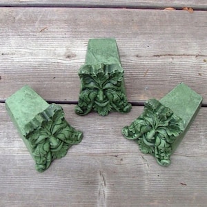May include: Three green concrete garden ornaments shaped like a face with leaves. The face is a traditional green man design.