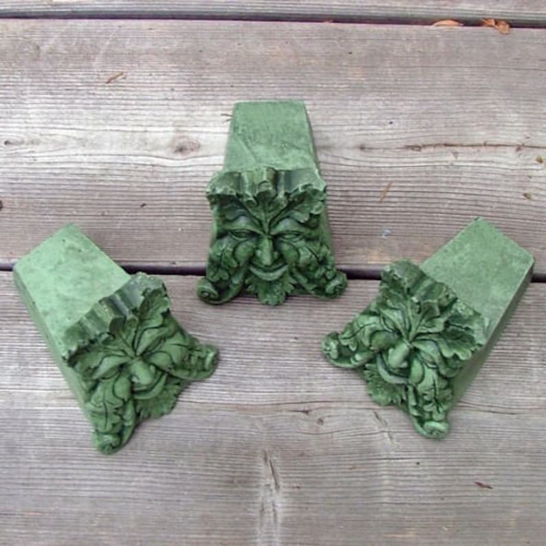 May include: Three green concrete garden ornaments shaped like a face with leaves. The face is a traditional green man design.
