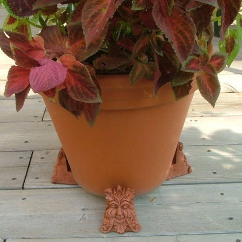 Op de afbeelding: Een terracotta plantenpot met een bladplant en decoratieve groene man voeten. De pot staat op een houten dek.
