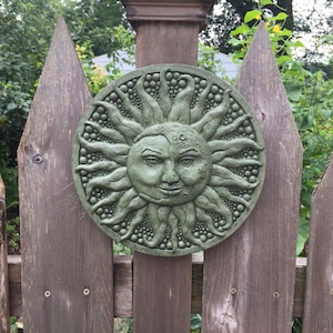 May include: A green ceramic sun and moon wall decor with a smiling face. The sun has a crescent moon shape on the right side of its face.