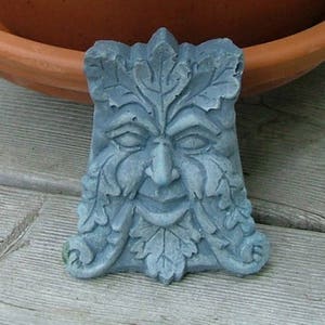 May include: A green and gray stone sculpture of a face with leaves. The face is smiling and has a beard made of leaves. The sculpture is a traditional green man design.
