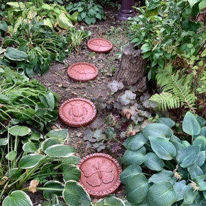 May include: Four terracotta stepping stones with different insect and butterfly designs. The stones are arranged in a path through a garden with lush green foliage.