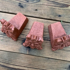 Op de afbeelding: Drie terracotta-kleurige keramische wigvormige blokken met een groen mannetje gezicht gegraveerd in de onderkant van elk blok.