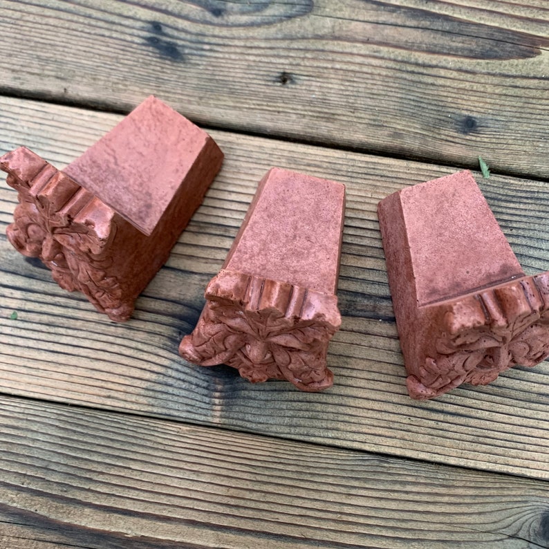 Op de afbeelding: Drie terracotta-kleurige keramische wigvormige blokken met een groen mannetje gezicht gegraveerd in de onderkant van elk blok.