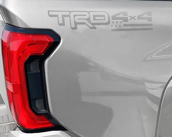 May include: Close-up of a silver vehicle's rear quarter panel. The image features a red taillight with a black border and the text "TRD 4x4" in gray. The design includes stars and stripes.