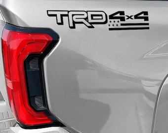 May include: A close-up of a silver vehicle's rear panel, featuring a black "TRD 4x4" decal. The decal includes the text "TRD" in a bold font, followed by "4x4" and a graphic of stars and stripes.