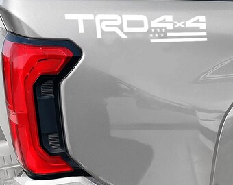 May include: A silver vehicle's rear quarter panel with a red taillight and white "TRD 4x4" decal. The decal includes stars and stripes, suggesting an American theme. The taillight has a black border.